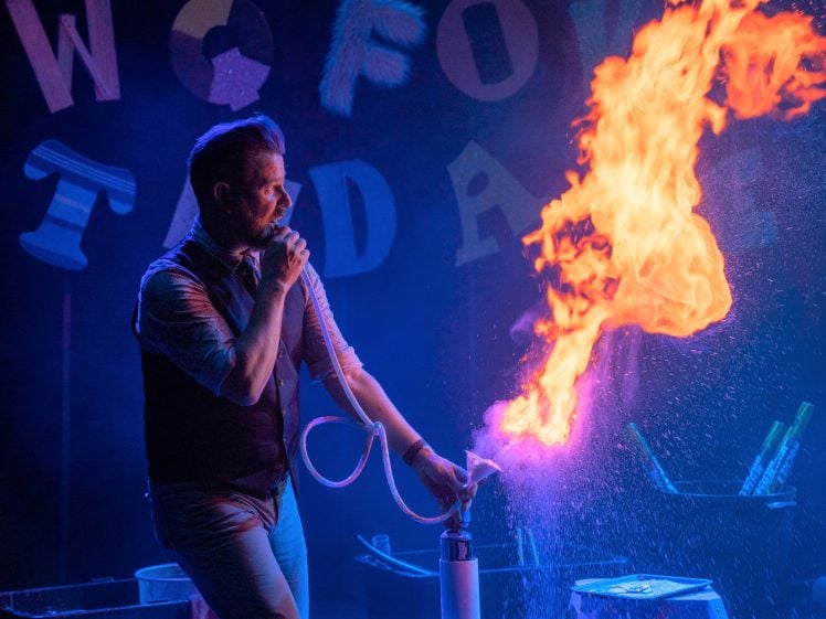 Image of A performer in a waistcoat creates a large burst of orange flames using a hose and spray bottle on a colourful stage with scattered letters in the background.