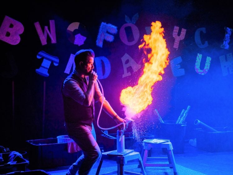 Image of A person creates a dramatic burst of fire using a tube and canister on stage, lit by vibrant blue and purple lighting, with large colourful letters hanging in the background.