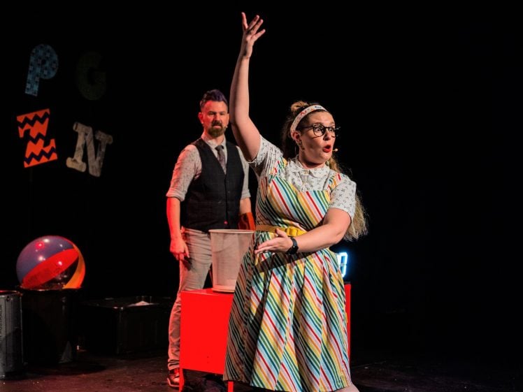 Image of A woman in a brightly striped dress gestures energetically on stage, while a man in a tie and waistcoat stands behind her. Colourful letters and a beach ball are visible against the dark background.
