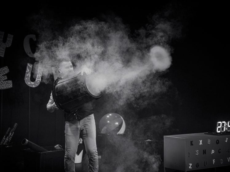 Image of A person aims a large cannon-like device that emits a thick cloud of smoke on a stage. Letters hang in the background, and a digital clock on a table shows “23:45.” The image is in black and white.
