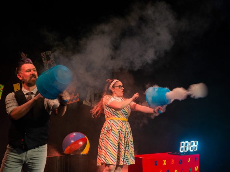 Image of Two people on stage, one man and one woman, shoot smoke from large blue cannons. The woman wears a colourful striped dress and sunglasses. There’s a timer, a beach ball, and letters in the background.