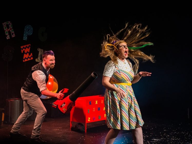 Image of A man uses a leaf blower to blow air at a surprised woman, causing her hair to fly wildly. Both are on stage with colourful props and letters in the background. The scene is playful and lively.