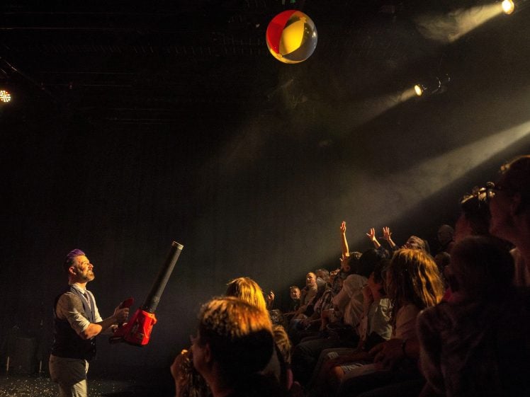 Image of A performer uses a leaf blower to launch a beach ball towards an enthusiastic audience, who reach up to catch it in a dimly lit theatre.