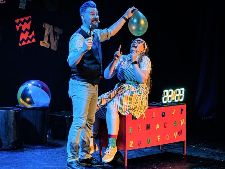 Image of A man holds a balloon and a pin near a laughing woman sitting on a colourful box with letters and numbers, on a stage decorated with letters and a beach ball. A digital clock displays 22:00 in the background.