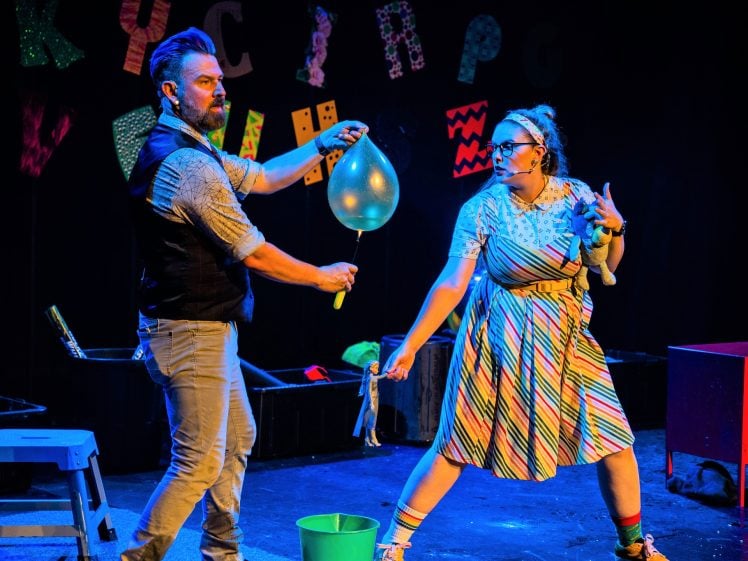 Image of A man holds a blue balloon with a stick whilst a woman in a striped dress reaches towards it on stage. Colourful letters are on the black backdrop, and props like a bucket and a box are nearby.