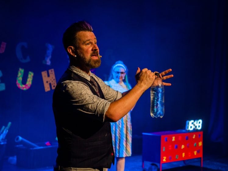 Image of A man with a headset microphone holds a clear bottle on stage, gesturing with his hand. A woman in colourful clothing stands behind him. Bright props and a digital clock display 16:49 are visible in the background.