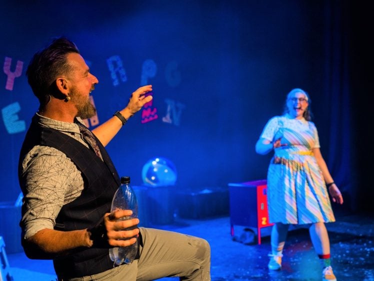 Image of Two people are performing enthusiastically on a colourful stage. The man in front holds a plastic bottle and gestures animatedly, while a woman in a striped dress stands in the background, reacting with surprise. Letters are hung as decorations behind them.