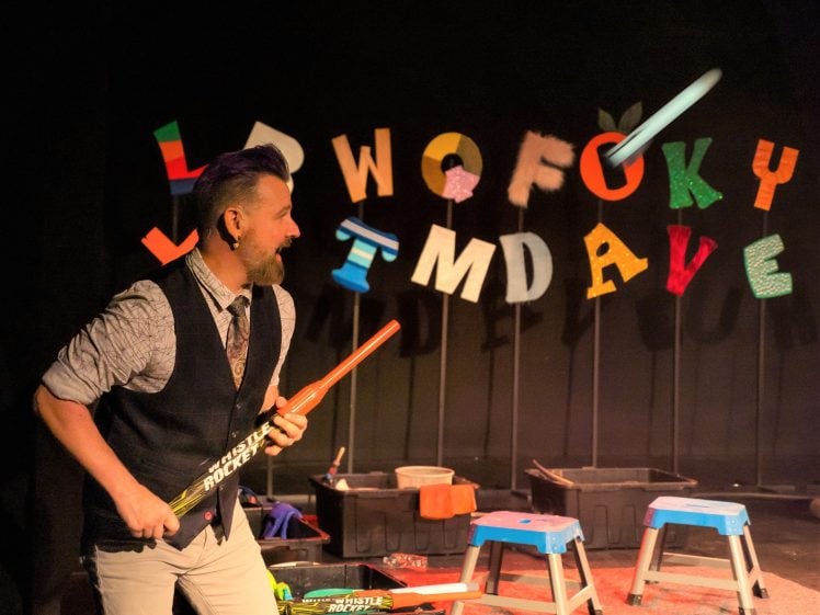 Image of A man with a beard and purple hair holds drumsticks on a stage, smiling. Behind him, colourful letters spell out LAWOFOKY TMDAVE on a black wall. Several stools and equipment are scattered about.