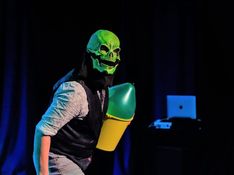 Image of A person wearing a bright green skull mask and holding a large green and yellow object stands on stage with dark curtains and electronic equipment in the background.