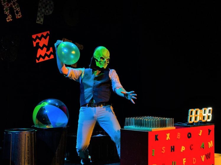 Image of A person wearing a bright green skull mask and waistcoat holds a green balloon on a dark stage with colourful props, including a beach ball, rubbish bins, letter board, and a digital timer reading 00:59.