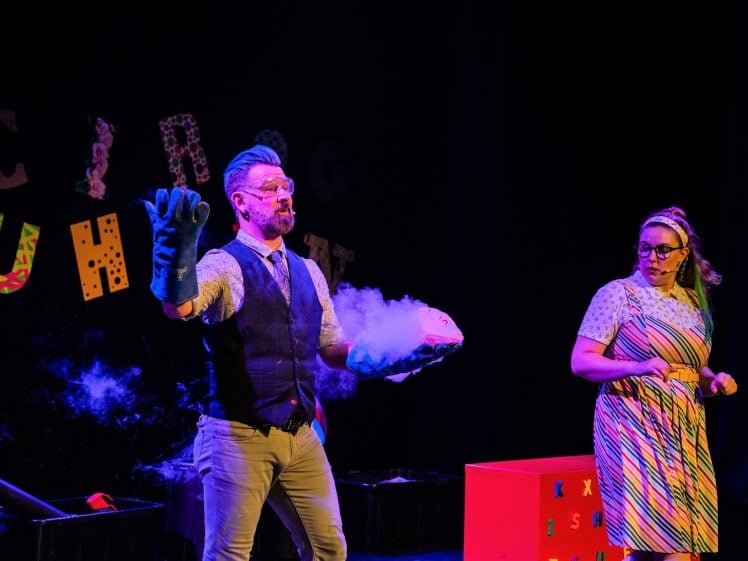 Image of A man wearing gloves and goggles holds a steaming bowl, with vapour rising, while a woman in a colourful dress and glasses watches beside him on stage. Brightly decorated letters and props are in the background.