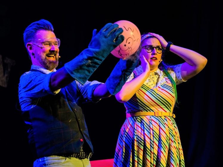 Image of A man wearing blue gloves and goggles holds a pink balloon with a cartoon face, while a woman in a colourful striped dress and glasses looks surprised, holding her head. Both are on stage with a dark background.