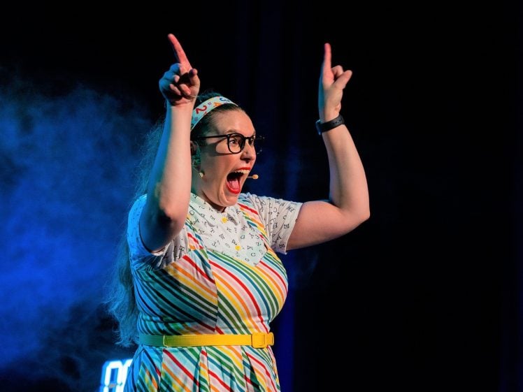 Image of A woman wearing glasses, a colourful striped dress, and a headset microphone is speaking energetically on stage, raising both arms with her index fingers pointed upward, with blue stage lighting and mist behind her.