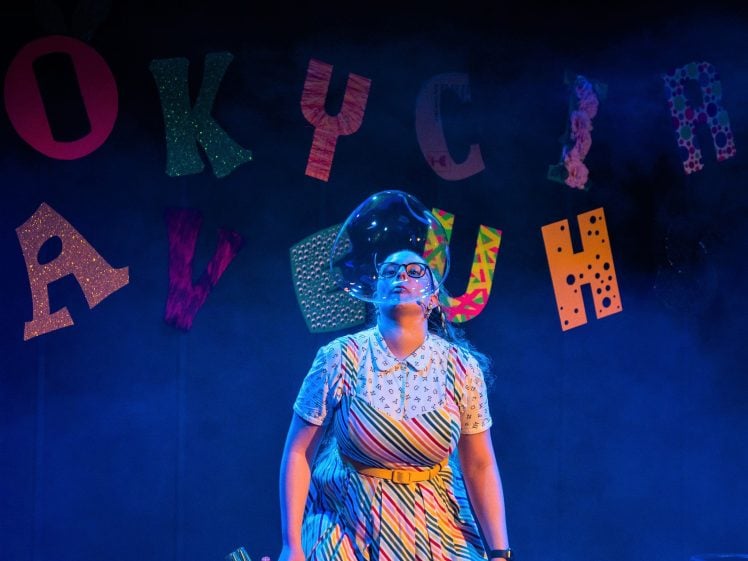 Image of A woman in a colourful striped dress stands on stage blowing a large bubble. Brightly coloured, patterned letters are arranged on the wall behind her, and blue stage lighting fills the scene.