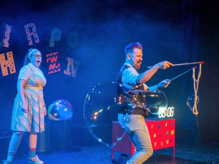Image of A man creates a large bubble with a rope wand on stage, whilst a woman in a striped dress and glasses watches excitedly. Colourful letters and a timer are visible in the background amid stage lighting and mist.