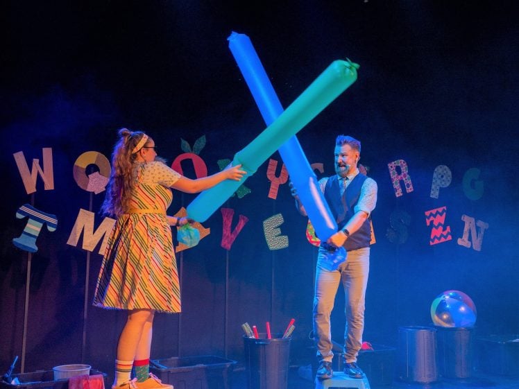 Image of A woman and a man playfully duel with large, blue foam batons on a stage decorated with colourful, cut-out letters and various props. The man stands on a small step ladder, and both are smiling.