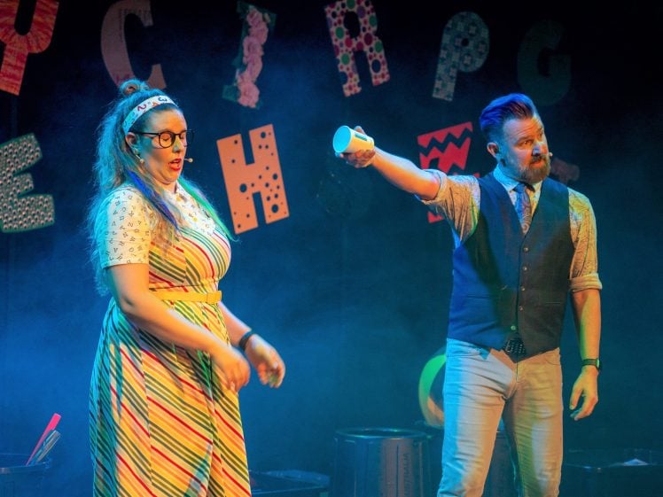 Image of Two performers on stage: a woman in a striped dress and glasses stands beside a man in a waistcoat holding up a white cup. Colourful, patterned letters hang in the background with blue stage lighting and smoke effects.