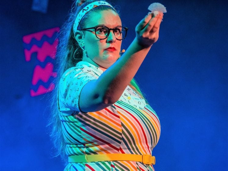 Image of A woman in a colourful, striped dress and glasses holds up a small, round object on stage, with blue lighting and abstract decorations in the background.