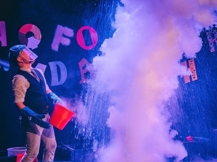 Image of A person wearing safety gear pours liquid from a red bucket, creating a large white cloud or splash on stage with colourful letters hanging in the background.