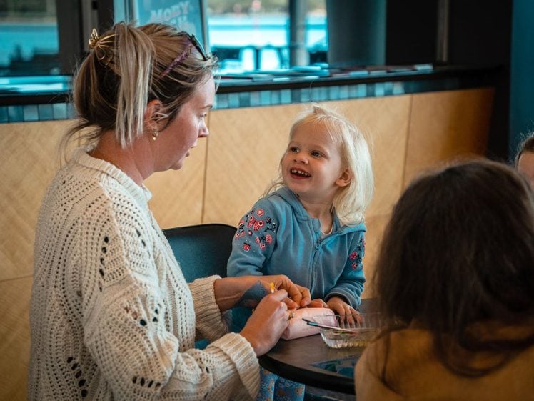 Image of A woman and a young girl sit at a table together. The woman holds the girl's hand while the girl smiles happily. Others are partially visible in the foreground, creating a warm, social scene indoors.