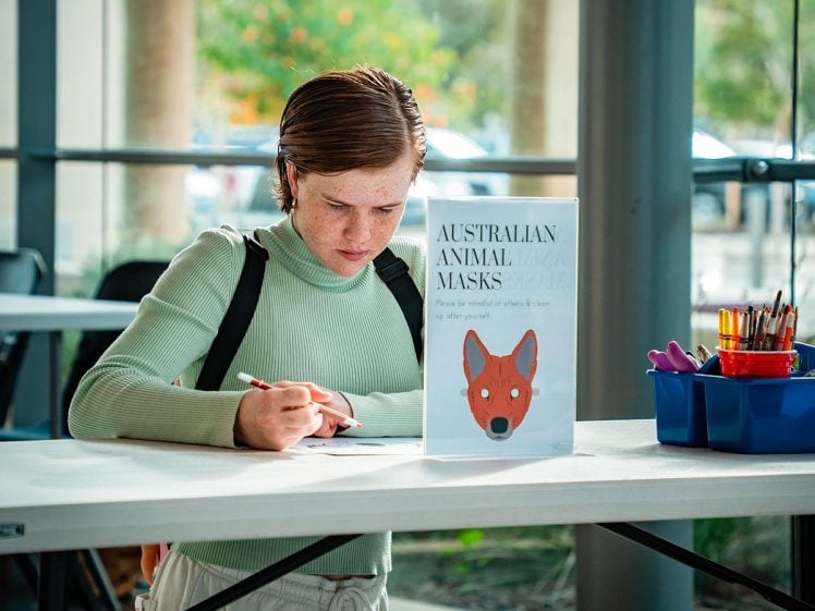 Image of A person with short brown hair in a light green top writes at a table. A sign on the table reads Australian Animal Masks with a drawing of a fox. Art supplies are in a blue caddy nearby.