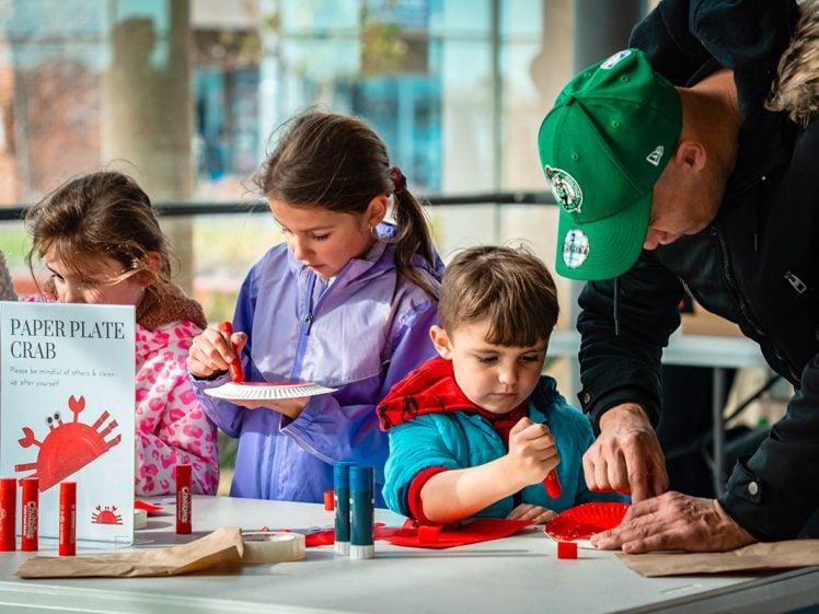Image of Four children and an adult work on a “paper plate crab” craft at a table, using red felt-tip pens and paper plates. A sign with instructions and a red crab illustration is displayed on the table.