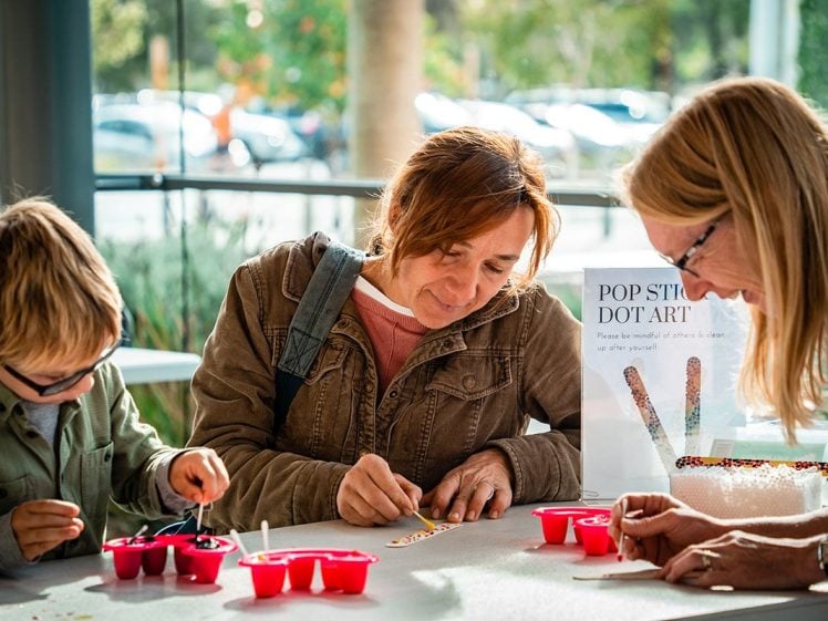 Image of A woman, a child, and another adult work on a craft project together at a table, using small colourful materials. A sign on the table reads POP STICK DOT ART. Sunlight streams through windows in the background.