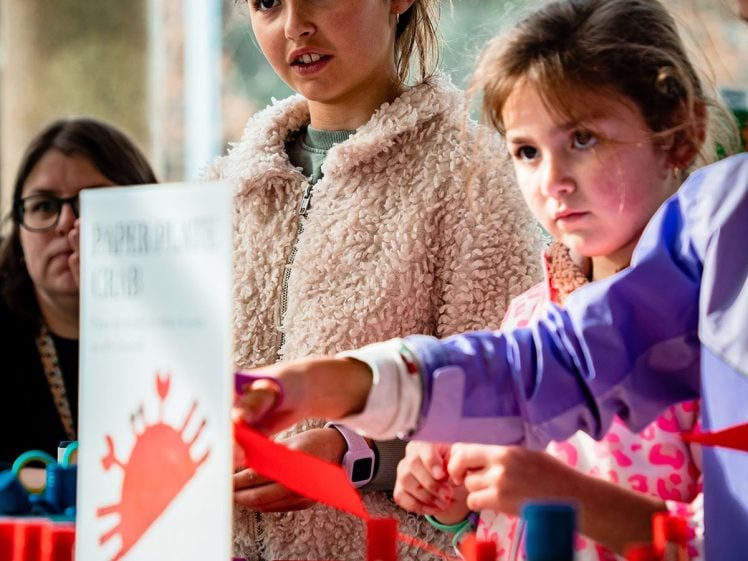 Image of Two young girls work together on a red paper craft project at a table, surrounded by art supplies. An adult and a craft instruction sign are visible in the background.