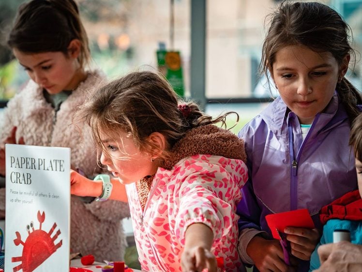 Image of Three young girls work on a “Paper Plate Crab” craft project at a table, focussing on their activities. A sign with crab artwork and crafting materials are visible in the foreground.