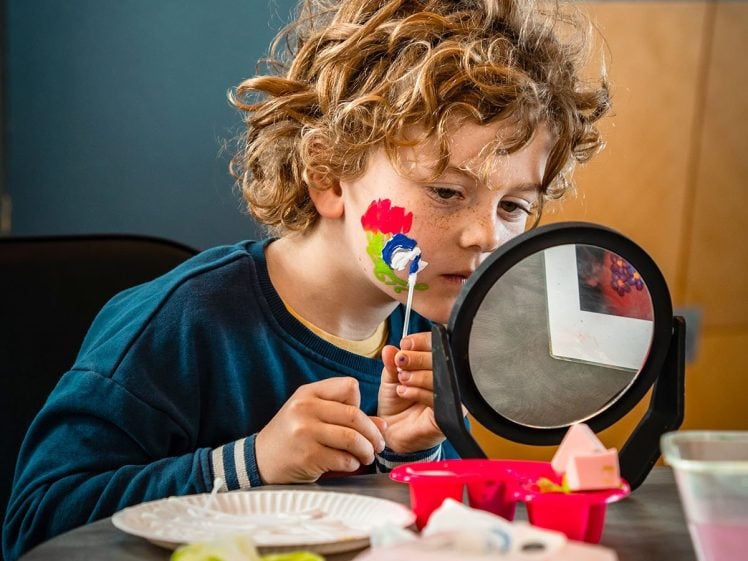 Image of A young child with curly hair paints a colourful flower design on their cheek while looking into a small mirror at a table covered with art supplies.