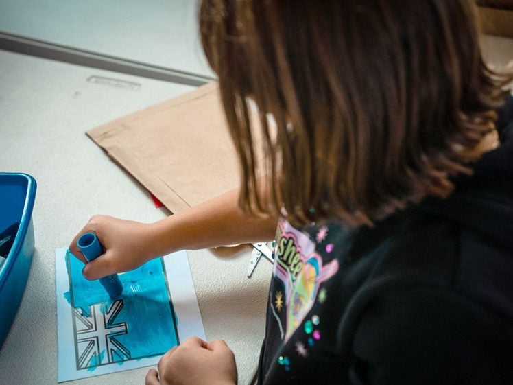 Image of A child is colouring a drawing of the Union Jack flag with a blue marker at a table. Art supplies are nearby and the child is wearing a black hoodie with a colourful design.