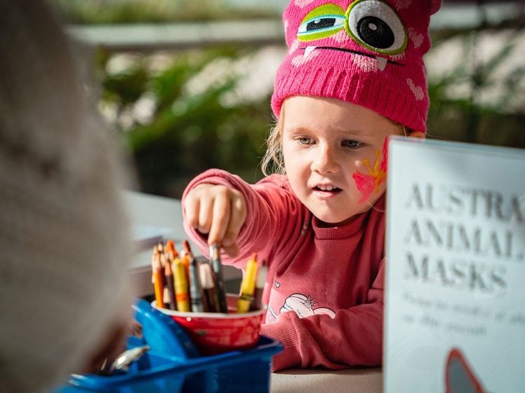 Image of A young child wearing a pink monster hat and face paint reaches for coloured pencils at a table, with a sign nearby that reads AUSTRALIAN ANIMAL MASKS.