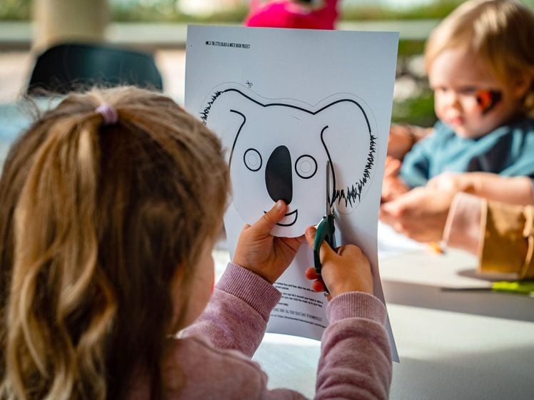 Image of A young child uses scissors to cut out a koala mask from a printed sheet, whilst another child with face paint sits in the background at a craft table.