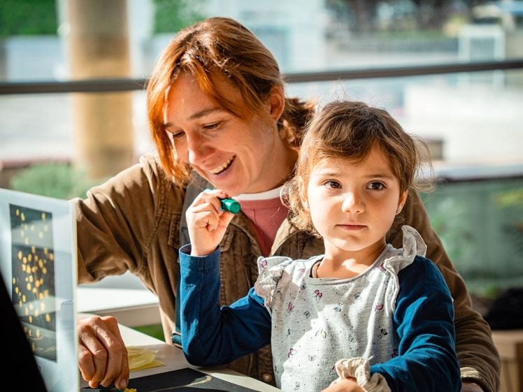 Image of A woman smiles whilst sitting beside a young girl who looks at the camera, holding a green object. They are indoors at a table with sunlight shining through large windows in the background.