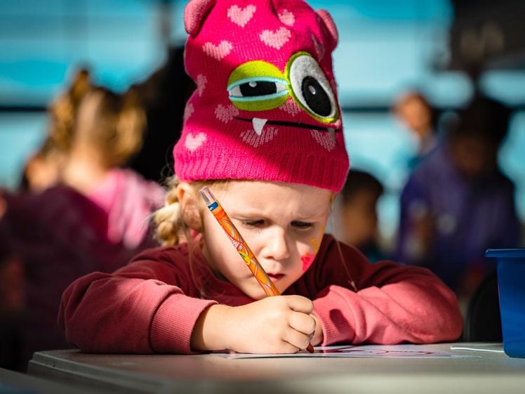 Image of A young child in a pink monster hat concentrates on drawing with a pencil at a table, surrounded by other children in a brightly lit room.