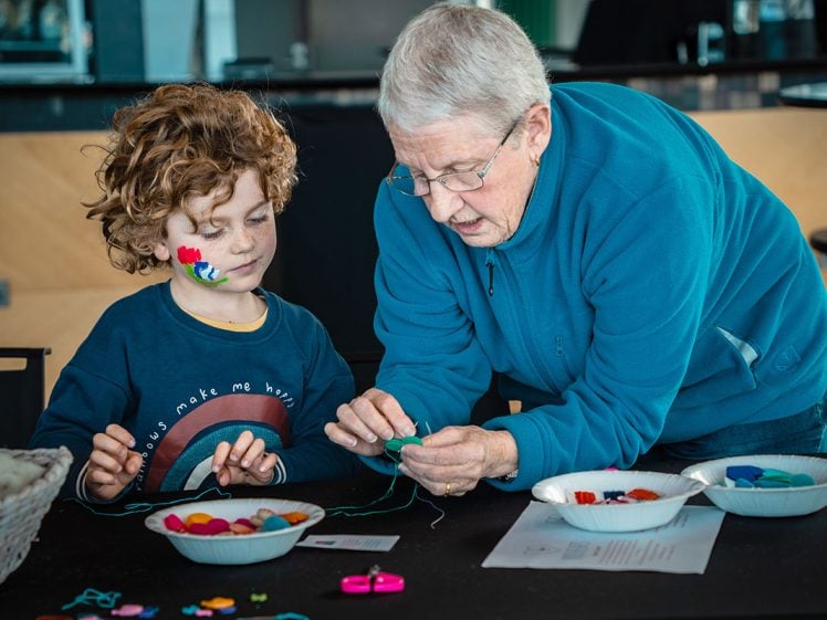 Image of An older woman helps a young child string colourful beads at a table. The child has a painted face and curly hair, and both are focused on the craft activity. Bowls of beads and supplies are on the table.