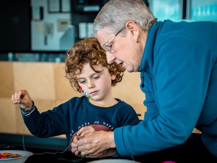 Image of An elderly woman helps a young child with curly hair thread beads onto a string. They are focussed on the craft activity, sitting together at a table in a bright, indoor setting.