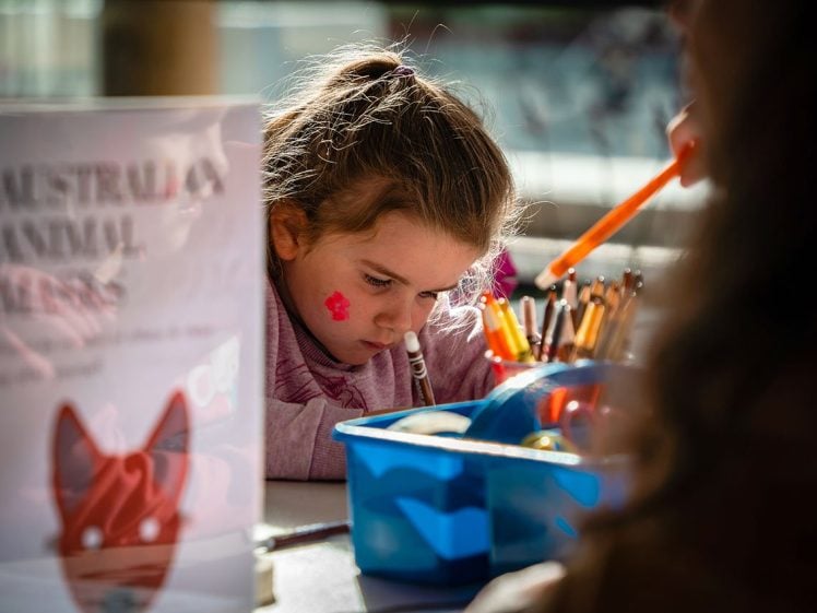 Image of A young girl with a pink flower painted on her cheek draws with coloured pens at a table, focused on her artwork. Sunlight shines on her face, and art supplies are scattered around. A sign about Australian animal masks is nearby.