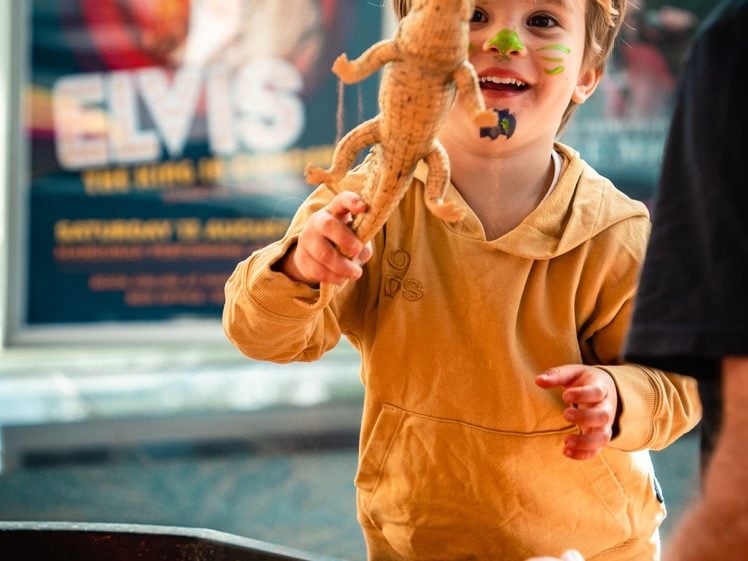 Image of A young child smiles and holds up a toy dinosaur whilst playing in a sandpit filled with toy animals. An adult’s hand is visible, and colourful posters are in the blurred background.