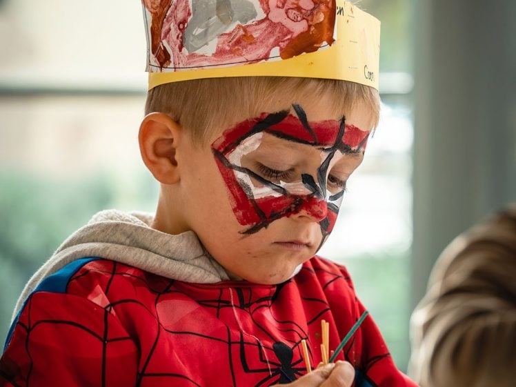 Image of Young child dressed in a Spider-Man costume, wearing a handmade paper crown and with their face painted like Spider-Man, focuses on painting or drawing at a table.