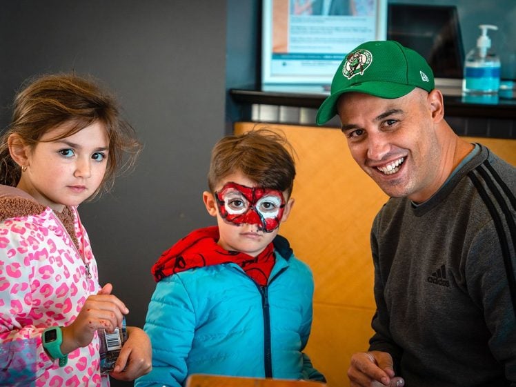 Image of A man in a green cap smiles next to two children; one wears a pink patterned jacket, and the other has a blue jacket and face paint resembling a superhero mask. They appear to be at a public indoor event.