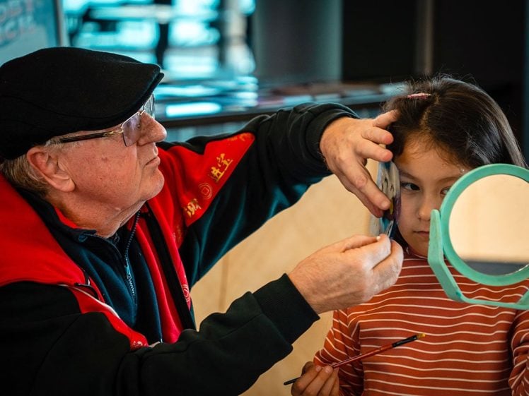 Image of An older man carefully paints the face of a young girl who is holding a round mirror and wearing a striped top. The man wears glasses, a black cap, and a red jacket.
