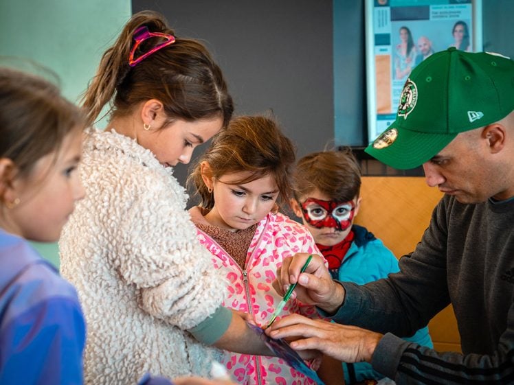 Image of A man in a green cap helps a group of children with an art or craft activity. One child has her arm held out, another looks on closely, and a child in the background has their face painted like a cat.