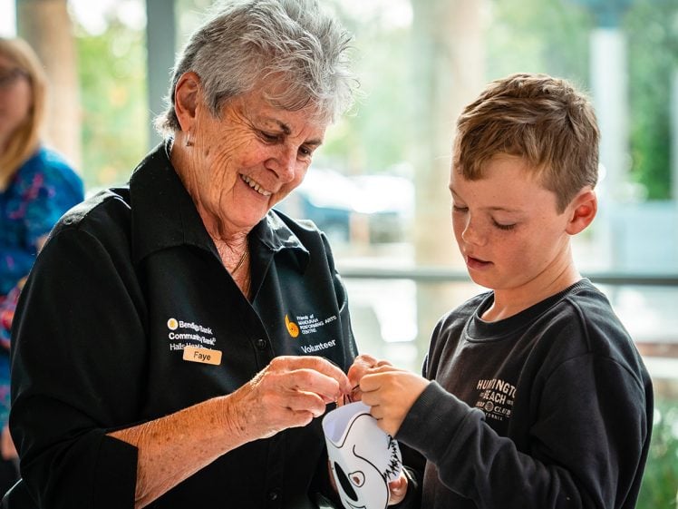 Image of An elderly woman and a young boy work together on a craft project, both smiling. The woman wears a black shirt with a volunteer badge, and the boy holds a paper cut-out shaped like an animal. Bright, blurred background.