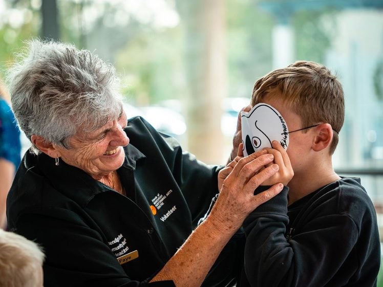 Image of An older woman smiles as she helps a boy put on a paper mask. The boy is laughing and holding the mask to his face. Both appear to be enjoying the playful moment indoors.