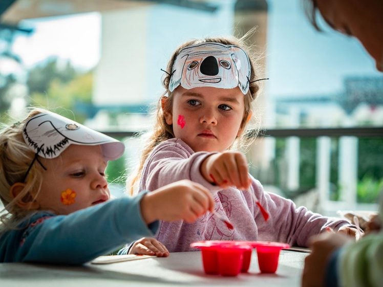 Image of Two young children wearing animal face masks and flower face paint sit at a table, engaged in an activity with red containers, whilst an adult assists them. The setting appears to be bright and outdoors.