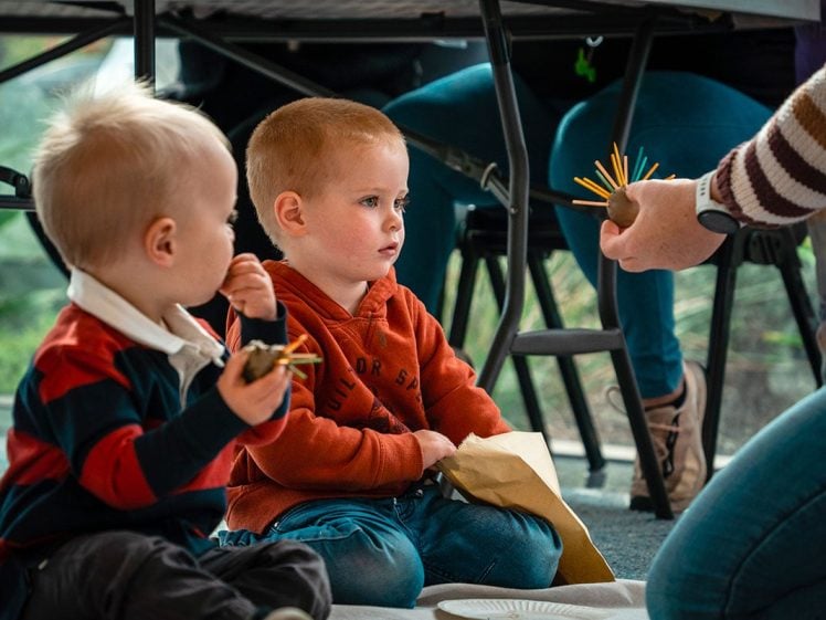 Image of Two young boys sit on the floor indoors, holding crafts. An adult extends a hand holding a craft made of sticks. Chairs and table legs are visible in the background. The atmosphere appears casual and focused.
