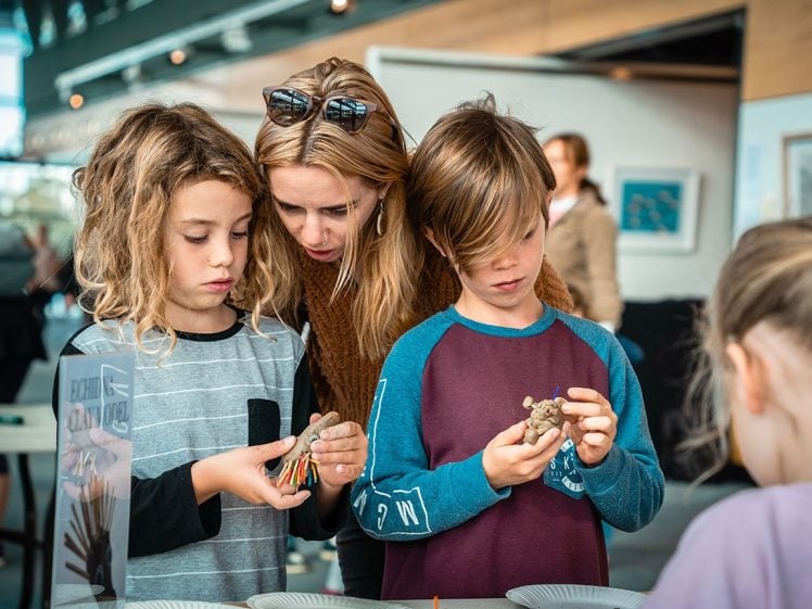 Image of A woman leans over two children as they closely examine small clay sculptures in their hands at a table, whilst another child sits nearby with her back to the camera in a bright indoor setting.
