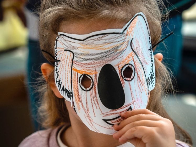 Image of A young child with light brown hair wears a hand-coloured koala mask, holding it to their face. The child is dressed in a pinkish sweatshirt with an illustrated character on it. The background is softly blurred.