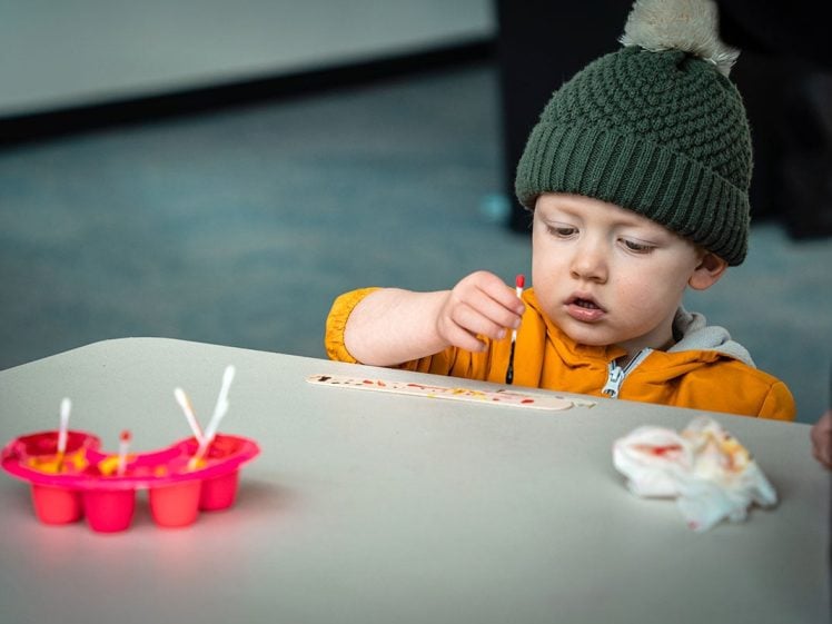 Image of A young child in a green knitted hat and yellow jacket sits at a table painting a wooden stick with a small brush. Art supplies and a serviette are on the table in front of them.